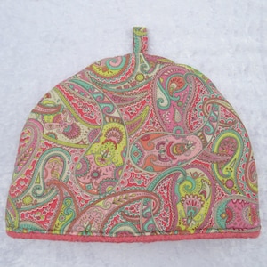 May include: A colorful tea cozy with a paisley pattern in shades of pink, yellow, and teal. The cozy has a loop at the top and a pink trim at the bottom. The tea cozy is designed to keep tea warm.