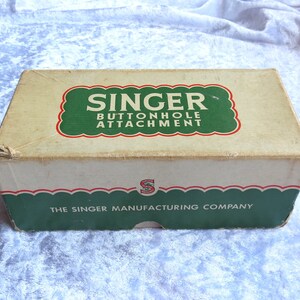 May include: A vintage Singer buttonhole attachment box with green and red accents. The box is labeled "Singer Buttonhole Attachment" and "The Singer Manufacturing Company".