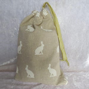 May include: A beige drawstring bag with a repeating pattern of white rabbit silhouettes and small white dots. The bag has a green and white checkered ribbon drawstring closure. The bag is made of a textured fabric.