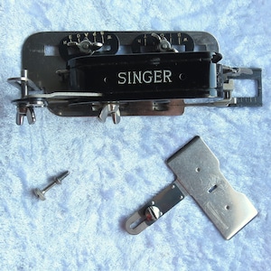 May include: A vintage Singer sewing machine attachment with a black metal body and silver metal accents. The attachment has a small ruler and a lever with a screw. The attachment is on a blue fuzzy surface.
