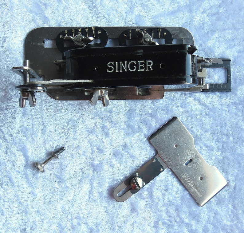 May include: A vintage Singer sewing machine attachment with a black metal body and silver metal accents. The attachment has a small ruler and a lever with a screw. The attachment is on a blue fuzzy surface.