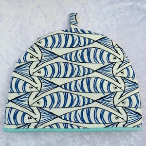May include: A tea cosy featuring a repeating pattern of blue and black fish on a white background. It has a light blue trim and a small loop at the top. The fish design is stylized, creating a nautical theme.