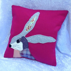 May include: A pink cushion with a grey and white rabbit applique. The rabbit has a floral patterned ear and a plaid patterned body.