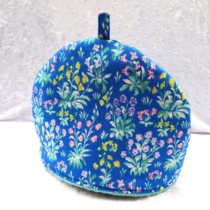 May include: A blue tea cosy with a floral pattern of pink, yellow, and purple flowers. The cosy has a loop at the top and a light blue trim. The floral design is set against a deep blue background. A kitchen textile.