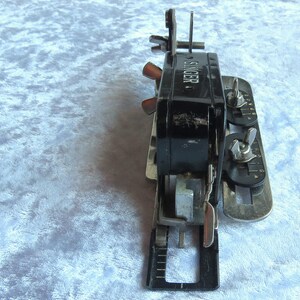May include: A black Singer sewing machine attachment with silver metal parts and a dial with numbers. The attachment has a lever and a knob.