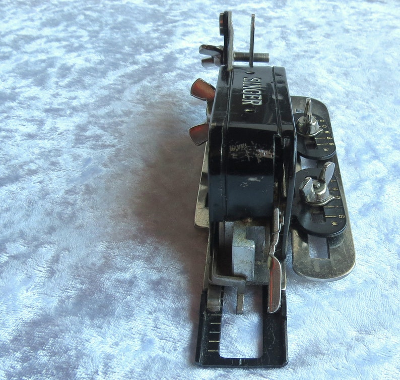 May include: A black Singer sewing machine attachment with silver metal parts and a dial with numbers. The attachment has a lever and a knob.
