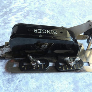 May include: A black Singer sewing machine attachment with a silver metal base. The attachment has a lever and two knobs labeled "Width" and "Space".