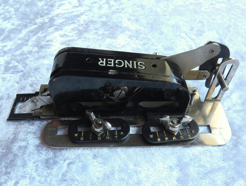May include: A black Singer sewing machine attachment with a silver metal base. The attachment has a lever and two knobs labeled "Width" and "Space".