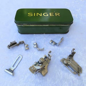 May include: A collection of vintage Singer sewing machine presser feet and accessories. The set includes various metal feet and a green tin box with "SINGER" in gold lettering on the lid. The items are arranged on a white surface.