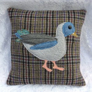May include: A gray and brown plaid cushion with a gray felt duck appliqué. The duck has a blue eye and a yellow beak.