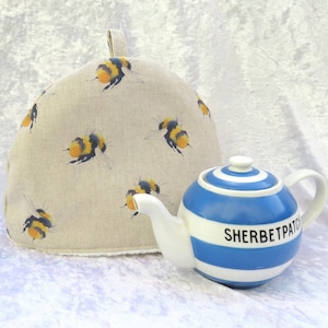 May include: A beige tea cosy decorated with yellow and black bumblebees. Next to it is a small, round teapot with blue and white stripes and the text "SHERBETPATCH" in black.