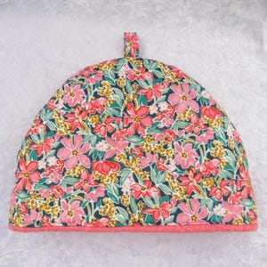 May include: A floral tea cozy with a pink trim. The cozy features a pattern of pink flowers, green leaves, and yellow accents on a dark blue background. It has a small loop at the top for easy handling.