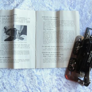 May include: A black Singer buttonhole sewing machine with a metal base and a white instruction booklet. The booklet has instructions on how to use the machine to create buttonholes of different lengths.