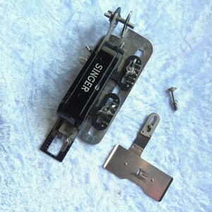 May include: A vintage Singer sewing machine attachment for creating buttonholes. The metal attachment has a black plastic component with the word "SINGER" printed on it. The attachment has several dials and knobs for adjusting the buttonhole size. There is a separate metal piece with a small screw.
