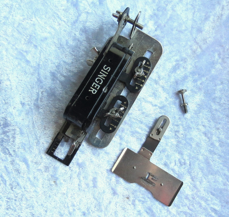 May include: A vintage Singer sewing machine attachment for creating buttonholes. The metal attachment has a black plastic component with the word "SINGER" printed on it. The attachment has several dials and knobs for adjusting the buttonhole size. There is a separate metal piece with a small screw.