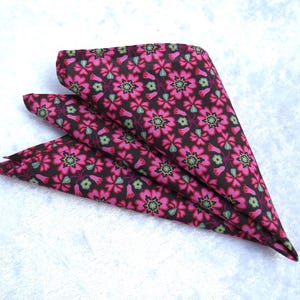 May include: Folded pocket square with a floral pattern. The fabric features a dark brown background with a repeating pattern of pink flowers with green and purple accents. The pocket square is folded into a triangle shape.