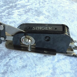 May include: Black Singer sewing machine attachment with a silver lever and a silver knob. The attachment has a black plastic body with the word "SINGER" embossed on it.