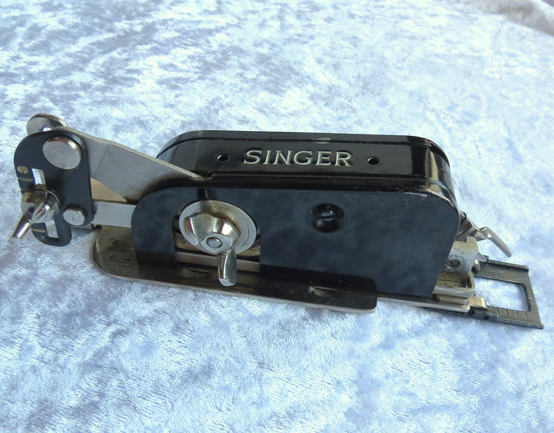 May include: Black Singer sewing machine attachment with a silver lever and a silver knob. The attachment has a black plastic body with the word "SINGER" embossed on it.