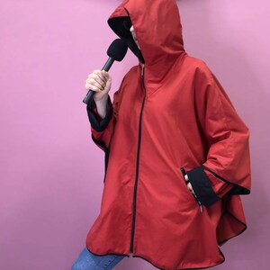 Puede incluir: Una chaqueta roja con capucha y ribetes negros y cierre de cremallera. La chaqueta tiene un estilo largo y fluido y está hecha de una tela ligera.