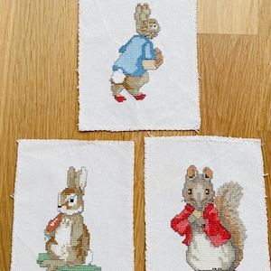 Puede incluir: Tres piezas bordadas de punto de cruz con Peter Rabbit, Benjamin Bunny y Squirrel Nutkin. Cada pieza está enmarcada con un borde de tela blanca.