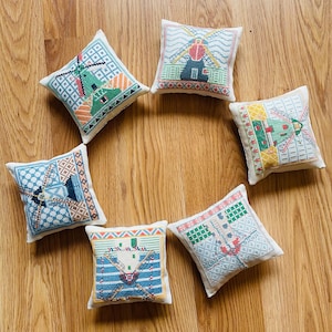 May include: Seven small, square, embroidered pillows with windmill designs in various colors, including blue, green, red, and yellow. The pillows are arranged in a circular pattern on a wooden surface.