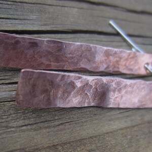 May include: A pair of hammered copper earrings with a textured surface. The earrings are a light copper color and have a rectangular shape.