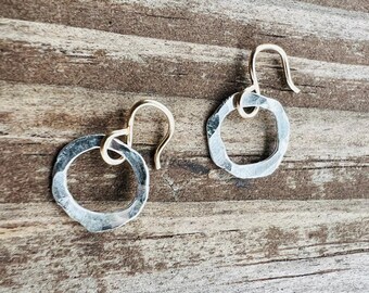 May include: A pair of silver earrings with a hammered texture and gold hooks. The earrings are circular in shape and have a rustic, handmade look.