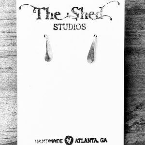 May include: A pair of silver teardrop earrings displayed on a white card. The card has the text "The Shed STUDIOS" at the top and "HANDMADE ATLANTA, GA" at the bottom. The earrings are hanging from the card.