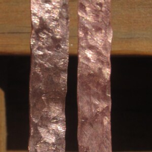 May include: A pair of hammered copper earrings with silver hooks. The earrings are a light pink color with a shimmery finish.