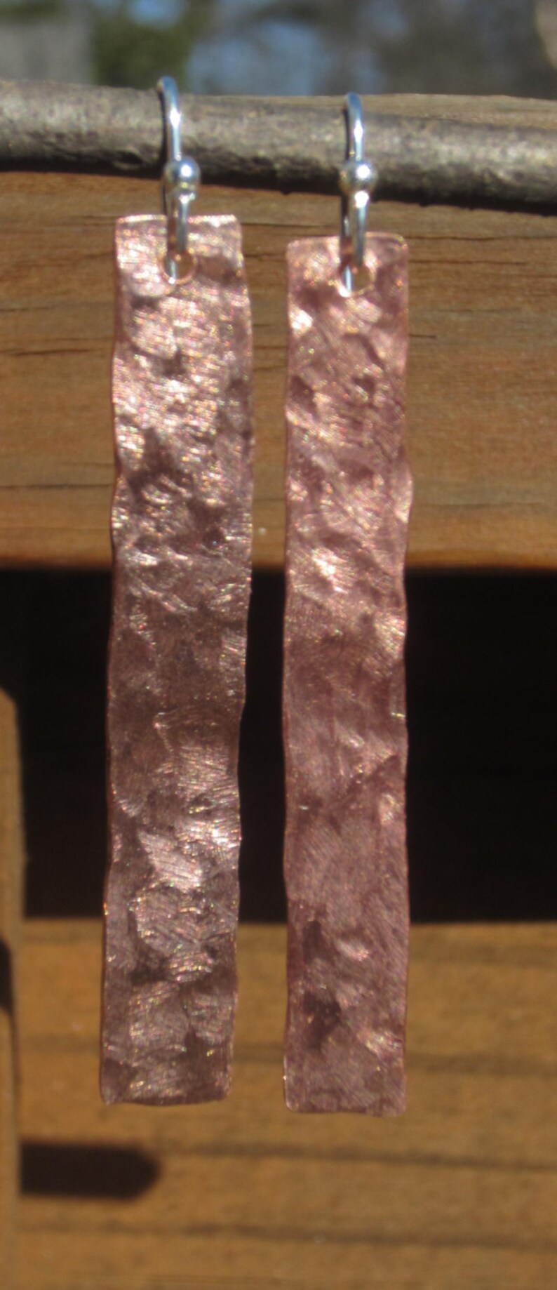 May include: A pair of hammered copper earrings with silver hooks. The earrings are a light pink color with a shimmery finish.