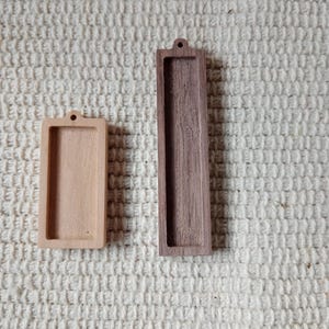 Op de afbeelding: Twee rechthoekige houten frames met een gat aan de bovenkant om op te hangen. Het frame aan de linkerkant is lichtbruin en het frame aan de rechterkant is donkerbruin.