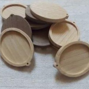 Puede incluir: Un conjunto de posavasos de madera sin terminar. Cada posavasos es redondo con un borde elevado y un pequeño agujero en un lado. La veta de la madera es visible, y los posavasos varían ligeramente en color, de marrón claro a medio.
