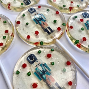 May include: Five clear round lollipops with a nutcracker image and red and green sprinkles. The lollipops are on white sticks.