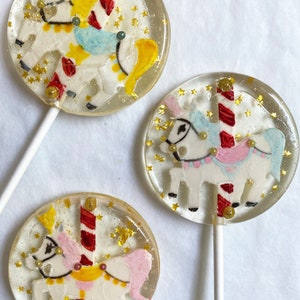 May include: Three clear lollipops with carousel horse designs. Each lollipop features a white horse with pink, blue, and yellow accents, surrounded by gold star-shaped sprinkles. The lollipops are on white sticks.
