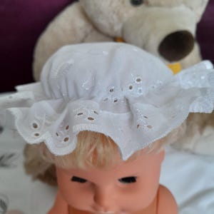Op de afbeelding: Een vintage pop met blond haar die een witte oogjesmuts draagt. De pop heeft een perzikkleurig gezicht en staat tegen een vage achtergrond met een teddybeer.