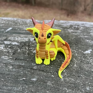 May include: A handcrafted, whimsical dragon figurine. The dragon features a yellow body with brown accents on its wings, horns, and spine. It has large, black eyes and is posed on a weathered wooden surface. The dragon is a fantasy-themed collectible.