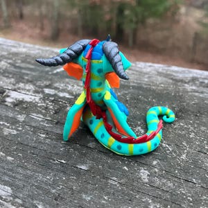 May include: A colorful, handcrafted dragon figurine. The sculpture features a turquoise body with yellow and blue accents, orange wings, and red spines. The dragon has gray horns and is posed on a wooden surface.