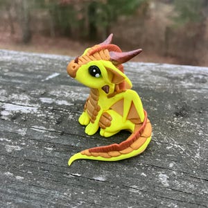 May include: A handcrafted, yellow and brown dragon figurine with large black eyes, horns, and wings. The dragon is made of clay and has a detailed, textured design. The figurine is sitting on a wooden surface.