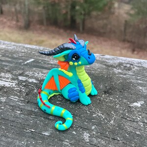 May include: A handcrafted, colorful dragon figurine. The sculpture features a teal body, blue head, and orange wings. The dragon has black horns and eyes, and is posed on a weathered wooden surface. A whimsical fantasy art piece.