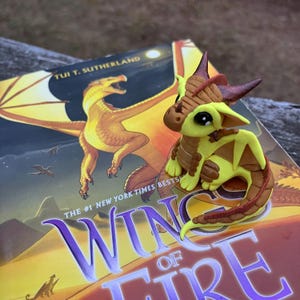 May include: A handcrafted polymer clay dragon figurine in shades of yellow, brown, and red, perched on a book titled "Wings of Fire" by Tui T. Sutherland. The book cover features a dragon illustration. The figurine has large black eyes and detailed wings.