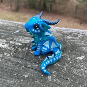 May include: A handcrafted blue and turquoise dragon figurine with white accents. The fantasy creature has horns, wings, and a long tail. The dragon is sitting on a weathered wooden surface.