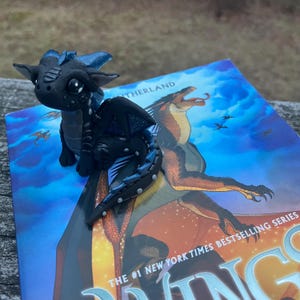 May include: A black and blue dragon figurine with large eyes sits on top of a book. The dragon has detailed wings and a textured body. The book cover features a dragon illustration and the text "Wings of Fire".