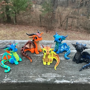 May include: Six colorful dragon figurines in various poses. The dragons are in shades of blue, orange, yellow, and black. Each dragon has horns, wings, and a long tail. They are displayed on a weathered wooden surface.