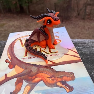 May include: A handcrafted orange and brown dragon figurine with black horns and large eyes, perched on a book titled "Wings of Fire: The Dragonet Prophecy". The book cover features a detailed illustration of a dragon.
