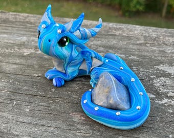 Op de afbeelding: Een handgemaakt blauw en zilveren drakenbeeldje met een edelsteen. De draak heeft grote groene ogen en is versierd met kleine, sprankelende versieringen. De sculptuur heeft een wervelend patroon van blauw en zilver en rust op een houten oppervlak.