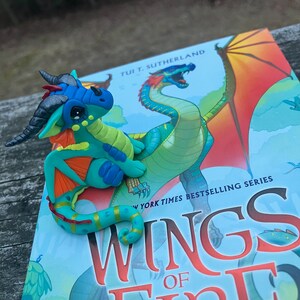May include: A handcrafted, colorful dragon figurine in shades of blue, green, and orange rests on a book. The book cover features a dragon illustration and the title "WINGS OF FIRE". The figurine has detailed features, including horns and wings.