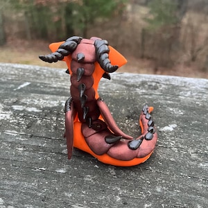 May include: A handcrafted orange and brown clay sculpture of a fantastical creature. The sculpture features dark brown horns, a segmented body, and black accents. The creature has a curved, tail-like form, and is set against a blurred outdoor background.