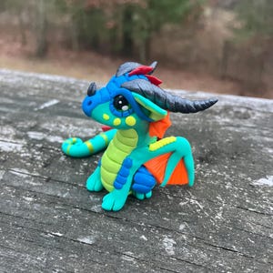 May include: A handcrafted polymer clay dragon figurine. The dragon features a blue head and body with green and yellow accents. It has orange wings and black horns. The dragon is posed on a weathered wooden surface.