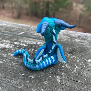 May include: A handcrafted blue and teal clay sculpture of a mythical creature with horns and a serpentine body. The sculpture features white accents and is posed on a wooden surface.