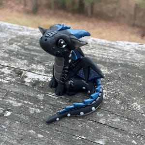 May include: A small, handcrafted dragon figurine in black and blue. The dragon has large, expressive eyes, detailed wings, and a long tail. The figurine is made of clay or a similar material and is posed sitting down.